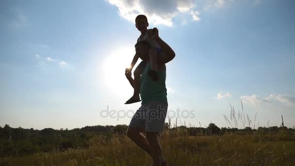 Ragazzino seduto sulle spalle del suo papà mentre cammina in un bellissimo paesaggio. Padre e figlio che giocano sul campo al tramonto. Famiglia felice passare del tempo all'aperto. Stile di vita attivo. Rallentatore — Video Stock