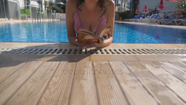 Retrato de menina bonita em chapéu e óculos de sol livro de leitura na piscina. Jovem relaxante no dia ensolarado quente durante as férias. Conceito de férias de verão. Vista frontal Fechar em câmera lenta — Vídeo de Stock