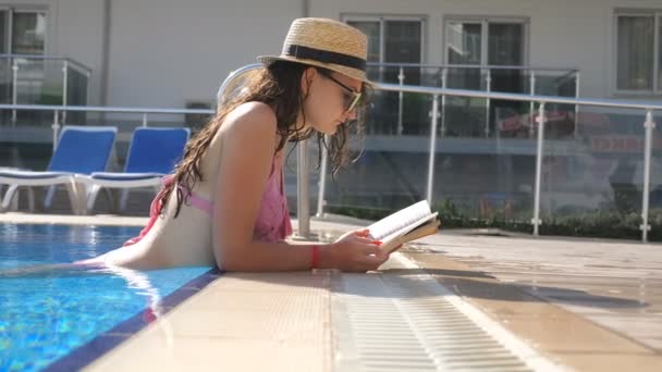 Krásná dívka v klobouku a sluneční brýle, čtení knihy v bazénu. Mladá žena uvolňující na teplý slunečný den během dovolené. Letní dovolená concept. Boční pohled zblízka zpomalené — Stock video