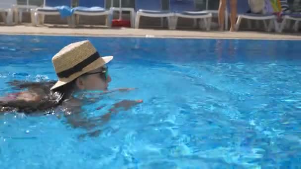 Menina bonita nova em óculos de sol e chapéu nadando na piscina. Mulher relaxando em água morna clara no dia ensolarado. Conceito de férias de verão ou férias. Fechar câmera lenta — Vídeo de Stock