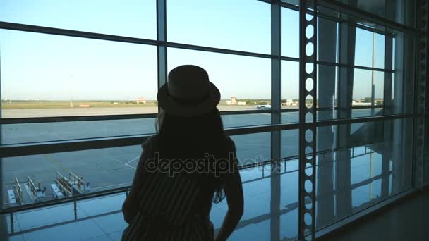 空港と滑走路を見てターミナルのウィンドウに来るバックパックと帽子の若い女。彼女の飛行機を待つ少女。観光と旅行のコンセプトです。背面バック見るスローモーション — ストック動画