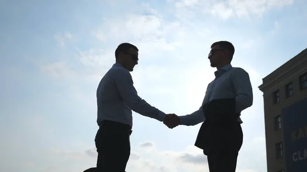 Dois jovens empresários que se encontram na cidade e se cumprimentam. Os colegas apertam as mãos ao ar livre. Aperto de mão de negócios ao ar livre. Agitação de braços masculinos lá fora. Céu azul com sol no fundo. Movimento lento — Fotografia de Stock