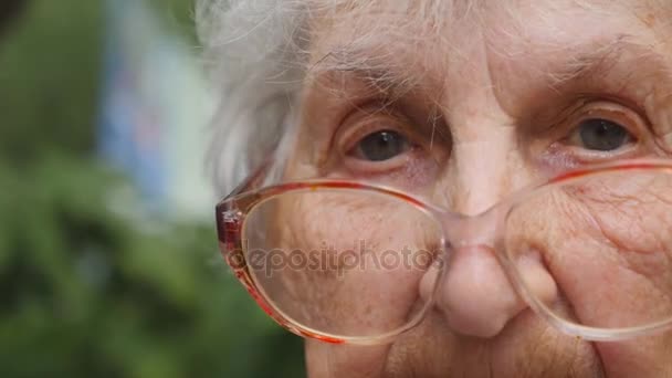 Старуха в очках поворачивает голову и смотрит в камеру. Бабушка в очках снаружи. Портрет бабушки на открытом воздухе. Закрыть Медленное движение — стоковое видео