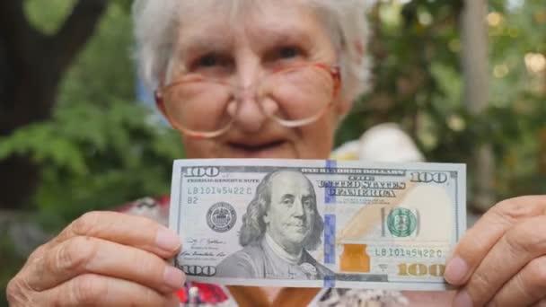 Ηλικιωμένη γυναίκα σε γυαλιά δείχνει εκατό δολαρίων νομοσχέδιο σε κάμερα και χαμογελαστός εξωτερική. Ευτυχισμένη γιαγιά κρατώντας ξένο νόμισμα εκτός. Έννοια του χρήματος. Εσωτερικη αργή κίνηση — Αρχείο Βίντεο