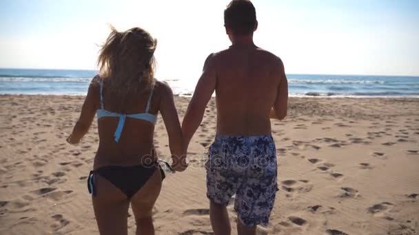 Молодая счастливая пара бежит на пляж к морю и держит друг друга за руки. Девочка и мальчик веселятся вместе на летних каникулах или каникулах. Закрыть задний обзор Медленное движение — стоковое видео