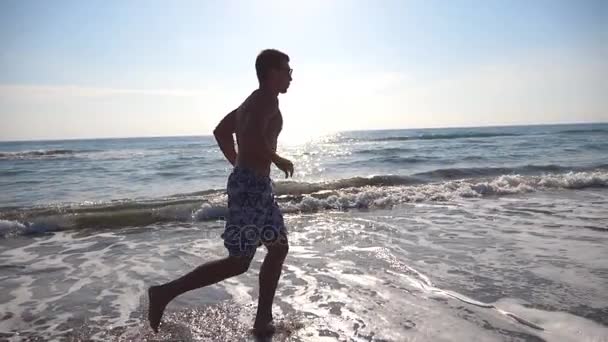 Ung sportig man kör på sea beach på morgonen. Atletisk kille jogging längs kusten på en solig dag. Manliga idrottsutövare tränar utomhus. Hälsosam aktiv livsstil. Sidovy Slow motion — Stockvideo