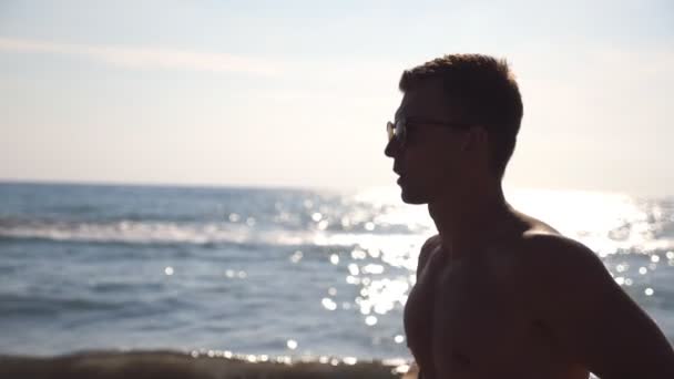 Profil sportovní chlap v sluneční brýle zaběhat podél pobřeží moře. Sportovní mladík běží za slunečného dne na pláži u oceánu. Mužské sportovce vykonávající venkovní. Zdravý aktivní životní styl. Detailní záběr Zpomalený pohyb — Stock video
