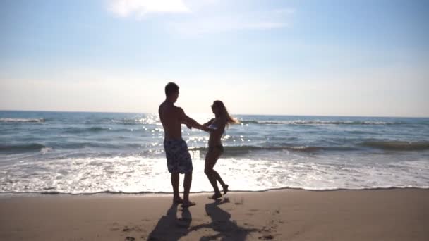 Ung man och kvinna höll händer och snurrar på havsstranden. Glada par virvlar runt tillsammans på en solig dag. Lyckliga par älskar att ha kul på stranden. Sommarsemester eller semester. Slow motion — Stockvideo