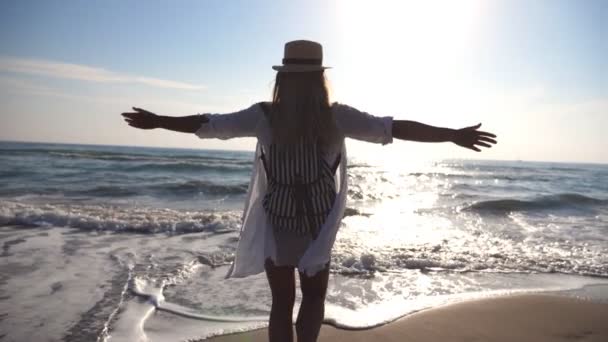 Mladá dívka v košili s batohem běhání k moři a zvyšování rukou. Šťastná žena v klobouku postřik vodou s nohama a užívat si života. Letní prázdniny nebo dovolenou. Pohled zezadu zpět Zpomalený pohyb — Stock video