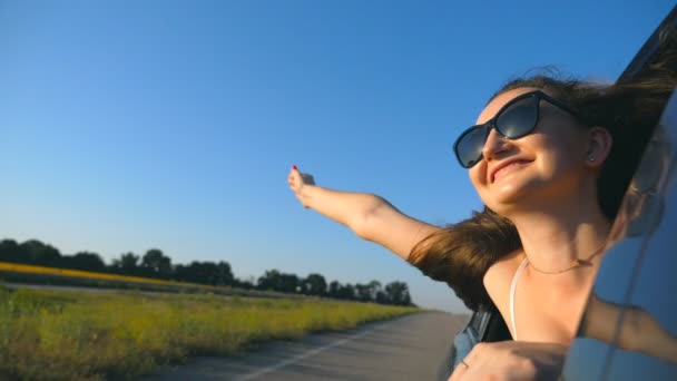 Chica feliz en gafas de sol que se inclina por la ventana del coche y disfrutar del viaje. Mujer joven mirando por la ventana de movimiento automático en el día soleado. Concepto de viaje y libertad. Cámara lenta Primer plano — Vídeos de Stock