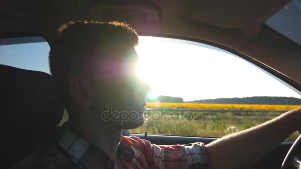 Profiel van:: jongeman in zonnebril besturen van een auto met zon flare op achtergrond. Knappe jongen auto rijden bij zonsondergang. Kaukasische jongen in zijn voertuig. Weergave van de kant van de salon. Slow motion close-up — Stockvideo