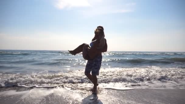 Happy man holding woman in his arms and spinning around on sea beach. Young guy circling his girlfriend in his hands on a sunny day. Couple in love spending time together during vacation. Slow motion — Stock Video