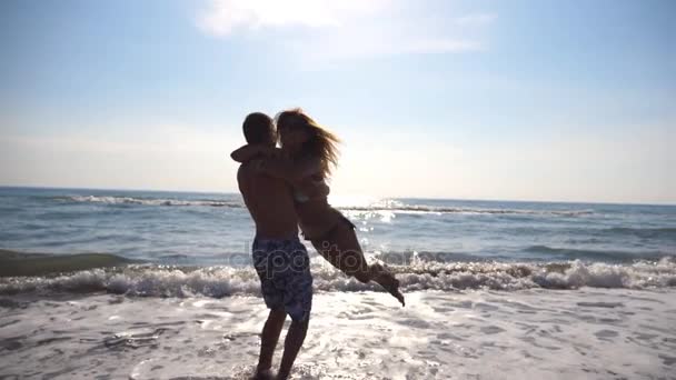 Giovane uomo che abbraccia e gira intorno alla sua donna sulla spiaggia. Fidanzato felice vorticoso fidanzata sulle sue mani a bella spiaggia. Gli amanti si divertono insieme in vacanza. Rallentatore Close up — Video Stock