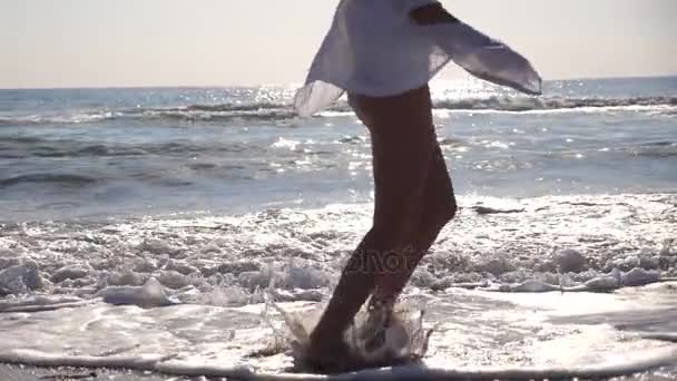 Πόδια γυναίκας με μπικίνι και πουκάμισο να τρέχουν στα κύματα της θάλασσας την αυγή. Ένα νεαρό όμορφο κορίτσι κάνει τζόκινγκ και διασκεδάζει στη θάλασσα. Θερινές διακοπές ή διακοπές. Κλείσιμο Πλευρική προβολή Αργή κίνηση — Αρχείο Βίντεο