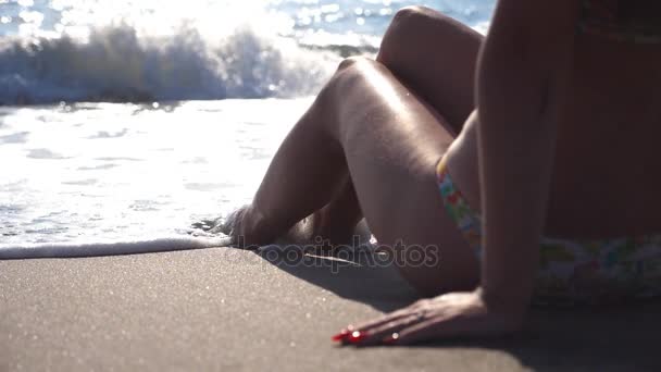 Ondas do mar a lavar-se sobre pés femininos bronzeados ao nascer do sol. Mulher bonita sentada na areia e relaxante perto da praia. Um pé de rapariga num resort. Férias de verão ou férias. Fechar câmera lenta — Vídeo de Stock