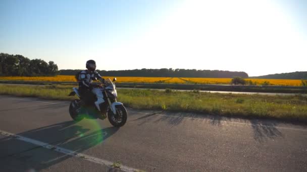 Motocyklista jazdy motocykl na drodze kraju z Słonecznikowe pole w tle. Młody człowiek w kasku jazda na rowerze moto o zachodzie słońca. Slow motion z bliska — Wideo stockowe