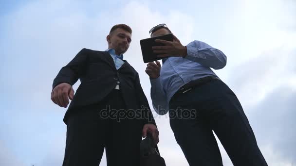 Два молодых бизнесмена разговаривают и используют планшетный компьютер на открытом воздухе. Деловые люди, работающие на цифровой планшет снаружи с небом на заднем плане. Коллеги применяют мобильные технологии. Низкий угол обзора — стоковое видео