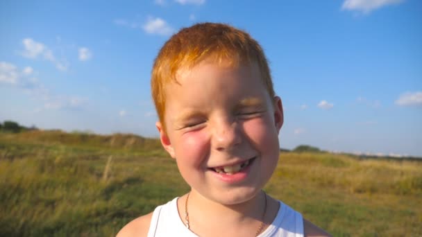 Gros plan sur les émotions de l'enfant mâle avec une expression heureuse sur le visage. Portrait de garçon aux cheveux roux heureux avec des taches de rousseur rit en plein air. Adorable beau bébé regardant dans la caméra avec un sourire joyeux. Mouvement lent — Video