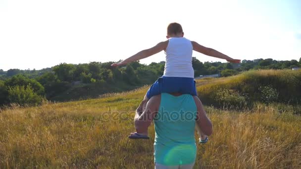 Vader en zoon spelen op het zomerveld op zonnige dag. Kleine jongen die op de schouders van zijn vader zit en de hand opsteekt als vliegtuig. Ouder met kind dat plezier heeft samen. Langzame beweging Achteruitkijkspiegel — Stockvideo