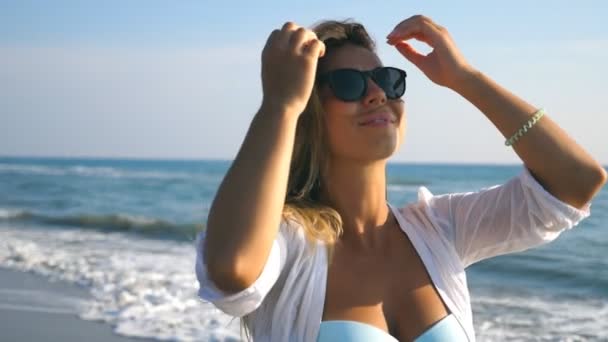 Nahaufnahme eines glücklichen Mädchens in Bikini und Hemd, das an der Küste entlang läuft und lächelt. junge schöne Frau mit Sonnenbrille, die mit ihren blonden Haaren spielt und im Sommerurlaub die Freiheit genießt. Zeitlupe — Stockvideo