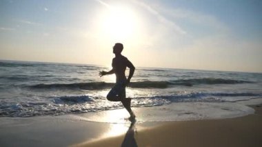 Hızlı deniz kıyı gündoğumu sırasında çalışan genç sportif adam silüeti. Atletik çocuk yalınayak kumsalda eğitim. Sığ sularda adım adam. Sağlıklı aktif yaşam tarzı. Ağır çekim yan görünüm