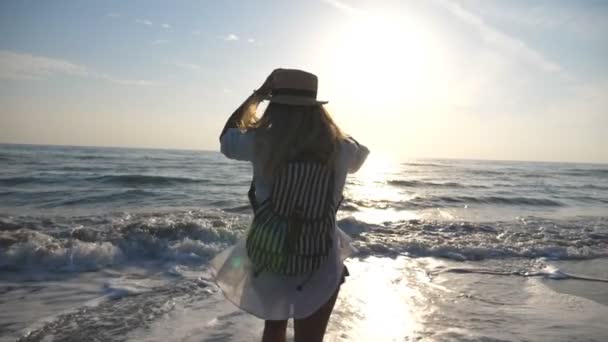 Ragazza felice in bikini e camicia con zaino che corre verso il mare e spruzza acqua con i piedi. Bella donna in cappello godendo la libertà e divertirsi all'aperto. Concetto di viaggio estivo. Rallentatore — Video Stock