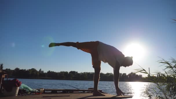 Silhuetten av ung sportig kille öva yoga flyttar och positioner i utkanten av träbryggan vid sjön. Atlet utbildning utomhus. Solljus på bakgrunden. Begreppet hälsosam aktiv livsstil. Närbild — Stockvideo