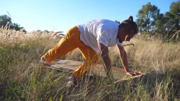 Молодой человек практикует движения йоги и позиции на ковриках на поле. Спортивный парень делает упражнения на лугу в солнечный день. Летний пейзаж на заднем плане. Концепция здорового активного образа жизни. Закрыть — стоковое видео
