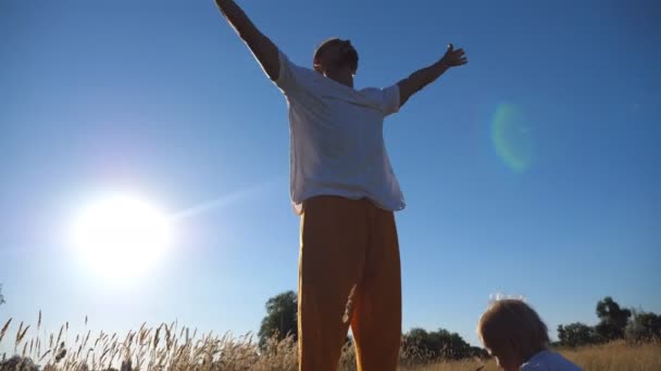 Malé dítě sedí na trávě na louce a jako jeho otec, cvičení jógy se pohybuje za slunečného dne. Mladý otec tráví čas se svým synem na pole. Sluneční světlo v pozadí. Detailní záběr pomalé mo — Stock video