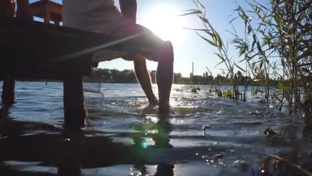 Nie do poznania człowiek siedzi na skraju drewniane molo nad jeziorem i wahadłowe nogi w wodzie. Młody facet, relaksujące i zwisające nogi w rzece w słoneczny dzień. Słońca światła w tle. Z bliska — Wideo stockowe