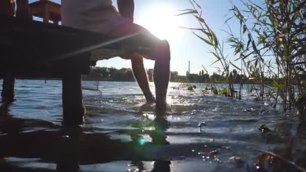 Unrecognizable man sitting on the edge of a wooden jetty at lake and swinging feet in the water. Young guy relaxing and dangling his legs in the river on a sunny day. Sun light at background. Close up — Stock Video