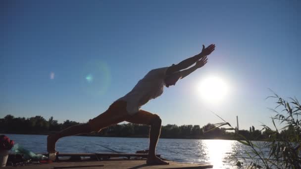 Silhueta de homem desportivo em pé na pose de ioga em um molhe de madeira no lago. Jovem a treinar na natureza. Atleta equilibrando em uma perna ao ar livre. Bela paisagem. Estilo de vida ativo saudável. Fechar — Vídeo de Stock