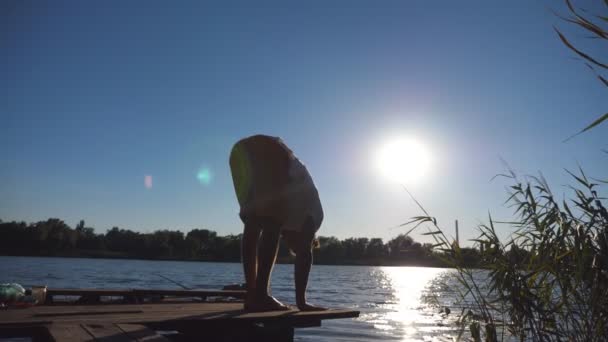 Jovem praticando movimentos de ioga e posições na borda de um molhe de madeira no lago. Atleta fazendo exercícios ao ar livre. Luz solar no fundo. Estilo de vida ativo saudável. Fechar para cima Visão traseira — Vídeo de Stock