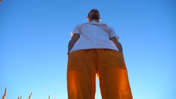 Low angle view of young man standing at the field and keeping his hands in pockets. Guy enjoying nature and freedom at the meadow on a sunny day. Blue sky at background. Close up — Stock Video