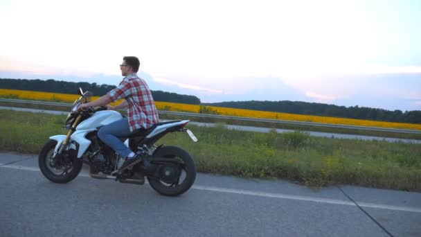 Young man riding on a modern sport motorbike. Handsome motorcyclist in a shirt and glasses driving his motorcycle on country road with sunflowers field at background. Slow motion Side view Close up — Stock Video