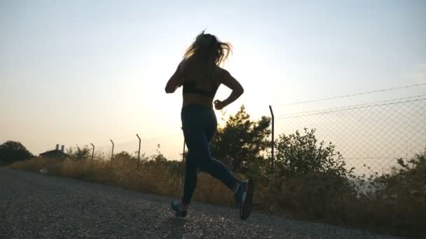 Följer till sportig flicka jogging på landet väg vid soluppgången. Ung kvinna springer utomhus på morgonen. En hälsosam aktiv livsstil. Långsamma rörelser — Stockvideo