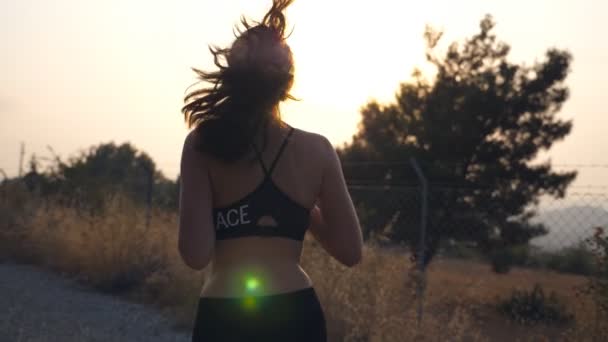 Follow to sporty girl jogging in country road. Young woman in sunglasses running outdoors and listening music from smartphone. Healthy active lifestyle. Slow motion Rear back view — Stock Video