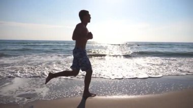 Deniz sahilde sabah koşan genç sportif adam. Sahil boyunca güneşli bir günde koşu atletik bir adam. Açık egzersiz erkek sporcu. Sağlıklı aktif yaşam tarzı. Yan görünümü ağır çekim