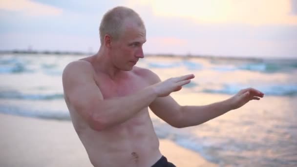 Mladý svalnatý muž cvičí boxerské cvičení na mořské pláži. Muž sportovec je cvičen v sebeobraně sám u pobřeží oceánu. Výcvik bojových umění pod širým nebem při západu slunce. Uzavření pomalého pohybu — Stock video