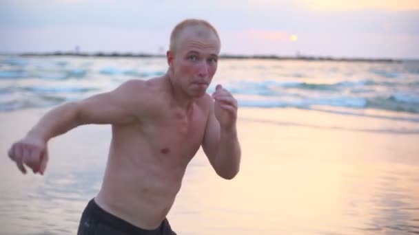Νεαρός μυώδης άνδρας που προπονείται σε ασκήσεις πυγμαχίας στη θάλασσα. Αρσενικό αθλητής ασκείται αυτοάμυνα μόνο κοντά στην ακτή του ωκεανού. Εκπαίδευση στις πολεμικές τέχνες υπαίθρια το ηλιοβασίλεμα. Αργή κίνηση Κλείσιμο — Αρχείο Βίντεο
