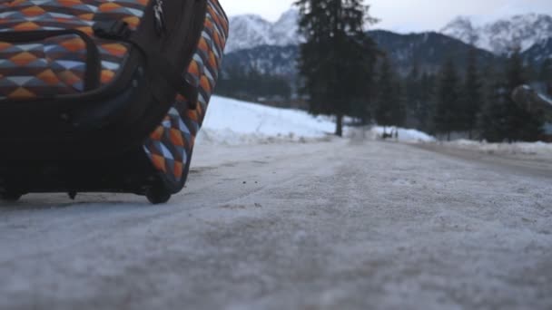 Hombre irreconocible que va con su equipaje en la carretera nevada durante el día de invierno. Un joven caminando por el sendero y rodando la maleta sobre ruedas. Paisaje de montaña al fondo. Vista trasera Vista lenta en cámara lenta — Vídeos de Stock