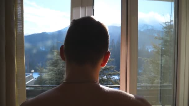 年轻的运动男子打开窗口在卧室的酒店和走出阳台在冬季的一天。英俊的家伙喜欢看山和森林在早晨。后后视图关闭慢动作 — 图库视频影像