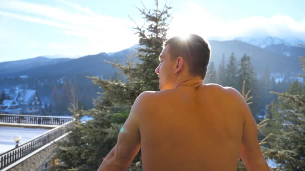 Primer plano del joven atlético parado en el balcón y respirando aire fresco durante el día de invierno. Hombre guapo disfrutando de hermosos paisajes de montaña y bosque por la mañana. Vista trasera Vista lenta en cámara lenta — Vídeos de Stock