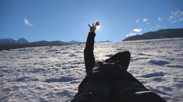 Gros plan d'un homme méconnaissable allongé sur la neige et regardant le soleil brillant à travers ses doigts par une journée ensoleillée. Main de jeune homme touchant la lumière du soleil sur fond de ciel bleu. Garçon heureux jouissant de la liberté — Video