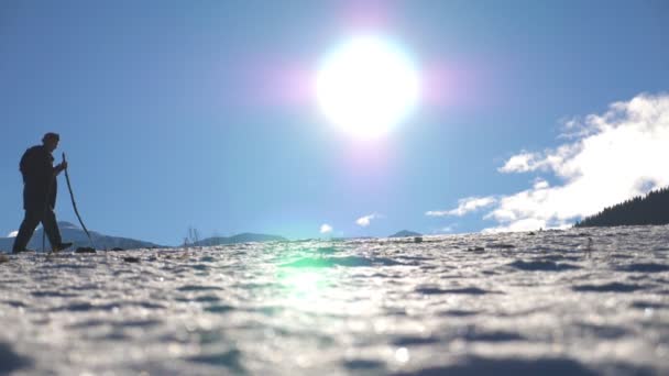 年轻的徒步旅行者, 北欧人带着棍子在太阳背景下漫步在雪地里。无法辨认的家伙在雪地上徒步, 在阳光明媚的日子在山上升起。健康的积极生活方式。侧面视图慢动作 — 图库视频影像