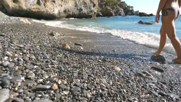 Vista trasera trasera de la niña caminando descalza en la costa en el día soleado. Suaves olas salpicando los pies de la joven mujer caminando en la playa de guijarros del mar. Concepto de vacaciones de verano o vacaciones. Movimiento lento — Vídeo de stock