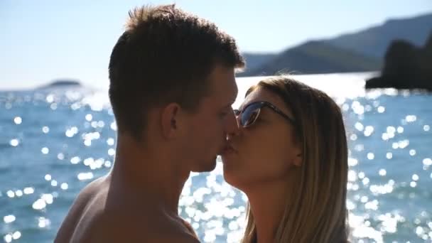 Close up of young couple kissing near seashore on sunny day. Beautiful pair spending time together on summer vacation. Sun light reflection at the sea water surface at background. Concept of loving — Stock Video