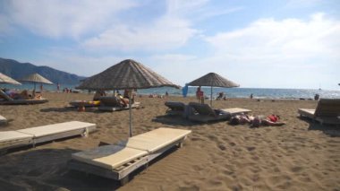 Dolly ile saman güneş şemsiyesi altında güverte sandalyeleri plaj güzel görünümüne güneşli bir günde bir kadeh. Güneşlenme yatakları sahil yakınındaki bronzlaşma ve yaz zevk tanınmaz turistler seyahat. Tatil kavramı