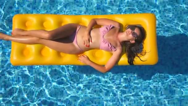 Mooi meisje in zonnebrillen en bikini, liggend op de gele opblaasbare matras in zwembad. Young gelooid vrouw die in de bekken van het hotel ontspannen op zonnige dag. Concept van de zomervakantie. Bovenaanzicht close-up — Stockvideo