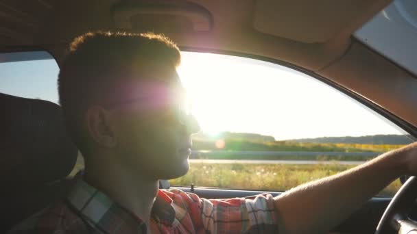 Profiel van:: jongeman in zonnebril besturen van een auto met zon flare op achtergrond. Knappe jongen auto rijden bij zonsondergang. Kaukasische jongen in zijn voertuig. Weergave van de kant van de salon. Slow motion close-up — Stockvideo